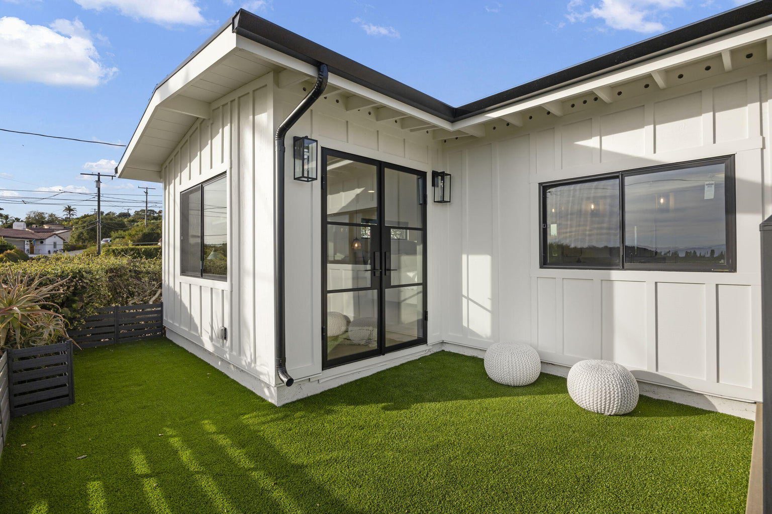 IMG_1189 A modern white exterior house with black trim, featuring a glass door that opens to a small yard covered with artificial grass and two round white outdoor stools.